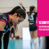Pallavolista con treccine e maglia nera e fucsia, a testa china sul campo da gioco, che esprime delusione dopo una sconfitta.